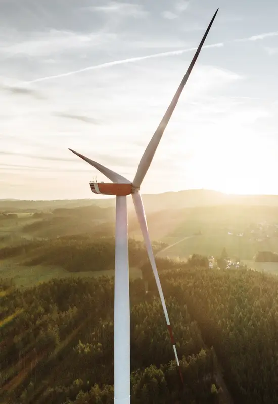 Aufwind – unser Newsletter für Grundeigentümer mit Weitblick Nahaufnahme eines Windrads bei Sonnenaufgang über bewaldetem Hügel (Hochformat).