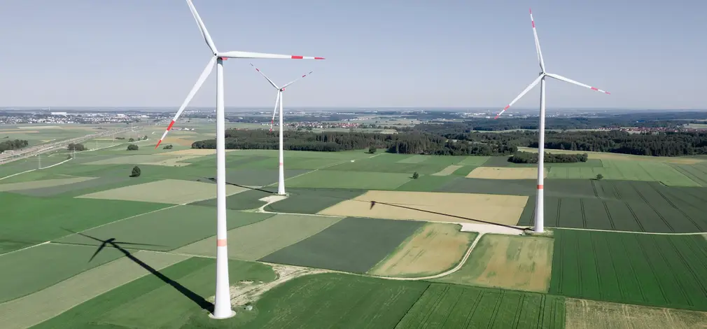Windpark mit weißen Windrädern über grünen Ackerfeldern und vereinzelten Bäumen unter hellem Himmel. Windpark mit weißen Windrädern über grünen Ackerfeldern und vereinzelten Bäumen unter hellem Himmel.