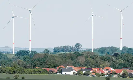 Windenergie in Kommunen – wie Städte und Gemeinden die Energiewende zum Heimspiel machen Mehrere Windräder stehen hinter einem Dorf mit roten Dächern und grünen Bäumen in ländlicher Landschaft.