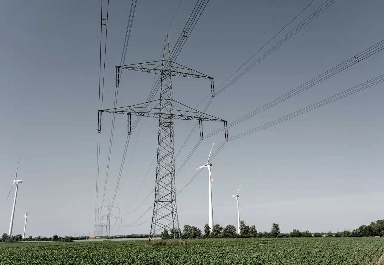 Deutschlands Zukunft in der Windenergie hängt davon ab, ob Ausbau, Netzanschluss, Betrieb und Lieferketten als durchgängiges System funktionieren. Windkraftanlagen und Hochspannungsleitung auf einem Feld.