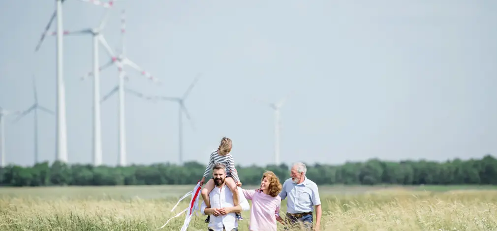 Familie mit Kind auf den Schultern läuft durch ein Feld, im Hintergrund mehrere Windräder und weiter Himmel.