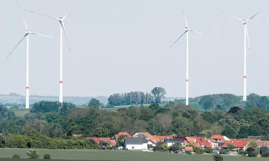 Windenergie in Kommunen – wie Städte und Gemeinden die Energiewende zum Heimspiel machen Mehrere Windräder stehen hinter einem Dorf mit roten Dächern und grünen Bäumen in ländlicher Landschaft.